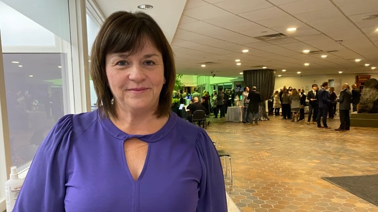 Pauline Fortin wearing a purple shirt. She is standing next to a window. Behind her on the other side there is a crowd of people at a reception.