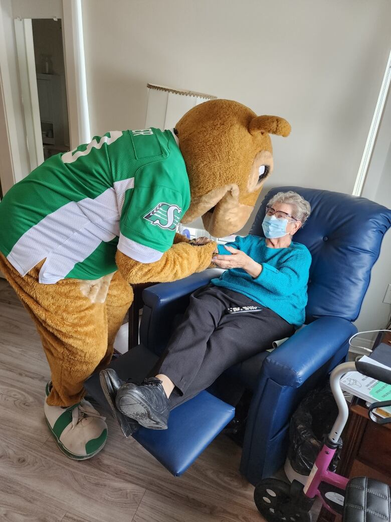 A mascot that is a squirrel wearing a Saskatchewan Roughriders jersey visits with an elderly woman who is seated on a recliner.