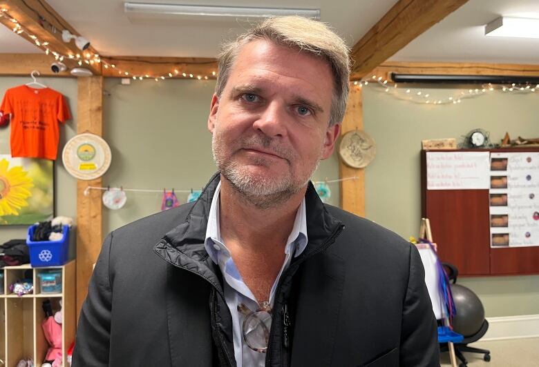 A man wearing a black jacket stands facing forward. His background is a classroom setting.