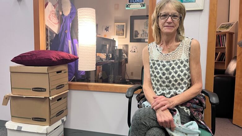 A woman sits on a chair in a lobby outside an office. Through the window behind her, you can see a desk, books and lamp. She's a middle-aged woman with blonde hair and glasses with a neutral expression.