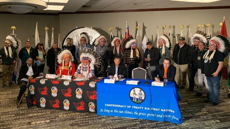 Four Treaty 6 chiefs, two wearing ceremonial headdresses, sit at a table while 16 other chiefs stand behind them in a hotel meeting room.