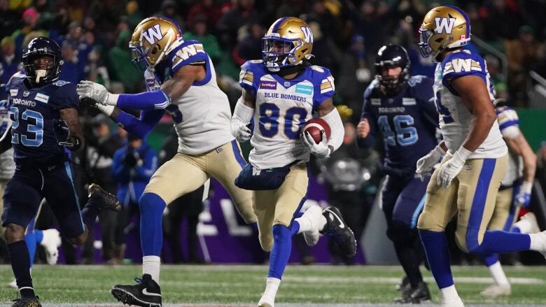 Winnipeg Blue Bombers team members run on green field wearing a gold, blue and white uniform. 