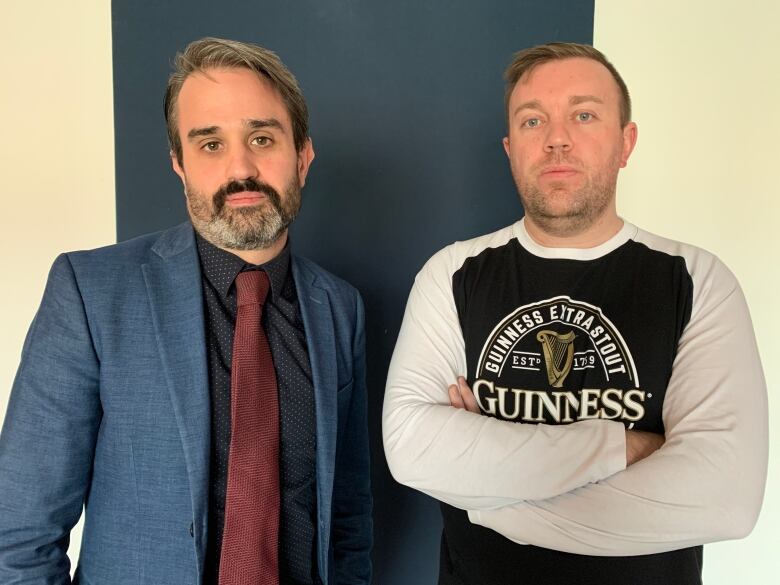 Two men standing against a wall, one with arms folded the other with arms at side. 