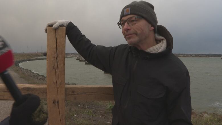 Man stands with a large wooden cross with a bridge in the background. 