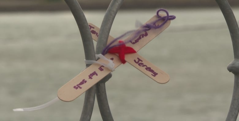 A small wooden cross with the name Jeremy written on it is attached to a metal bridge railing. 