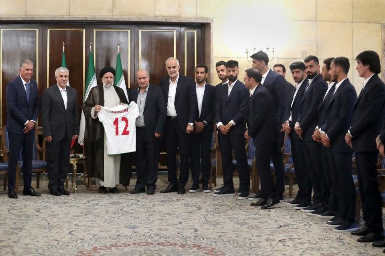 More than a dozen men in suits stand in a semi-circle with a man in long robes holding a soccer shirt with the number 12.