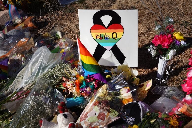 A pile of flowers, stuffed animals and rainbow flags on the ground surrounding a small handmade sign with the Cub Q logo: A rainbow heart over a black ribbon, with the words: 