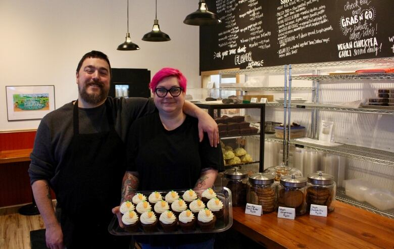 A man stands next to a woman, who is holding a tray of cupcakes. Both are smiling. They are standing in front of a bakery counter with filled display cases and cookie jars. A chalkboard on the wall behind them in scribbled with menu items.