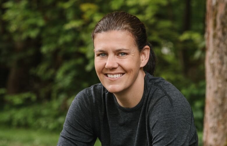 A woman wearing distressed jeans and a charcoal grey shirt smiles as she poses while sitting on a bench in front of a tree. 