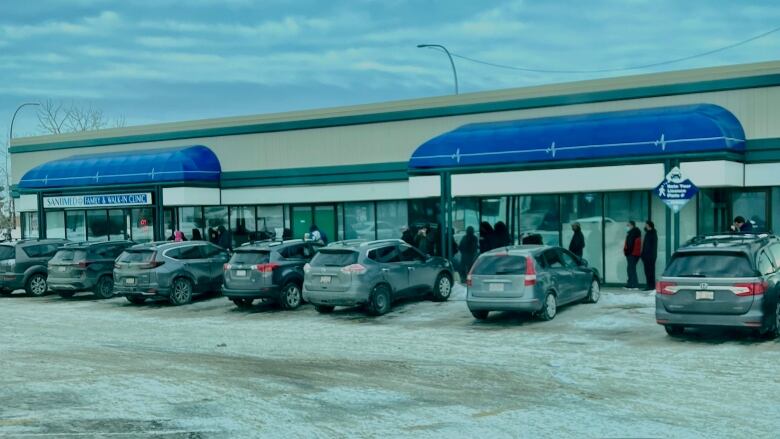 Vehicles sit in a snowy parking lot. Patients can be seen standing in line outside a clinic. 