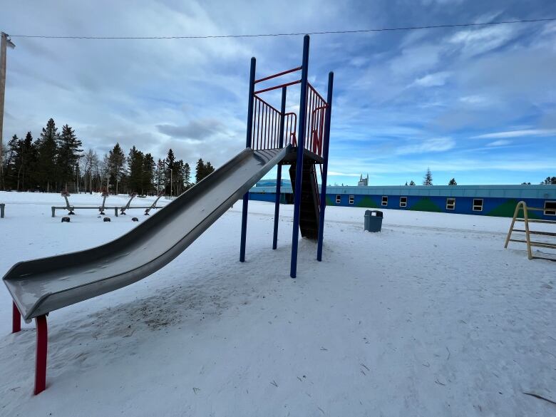 A school yard slide is seen in front of a low-lying elementary school painted in two tones of blue.