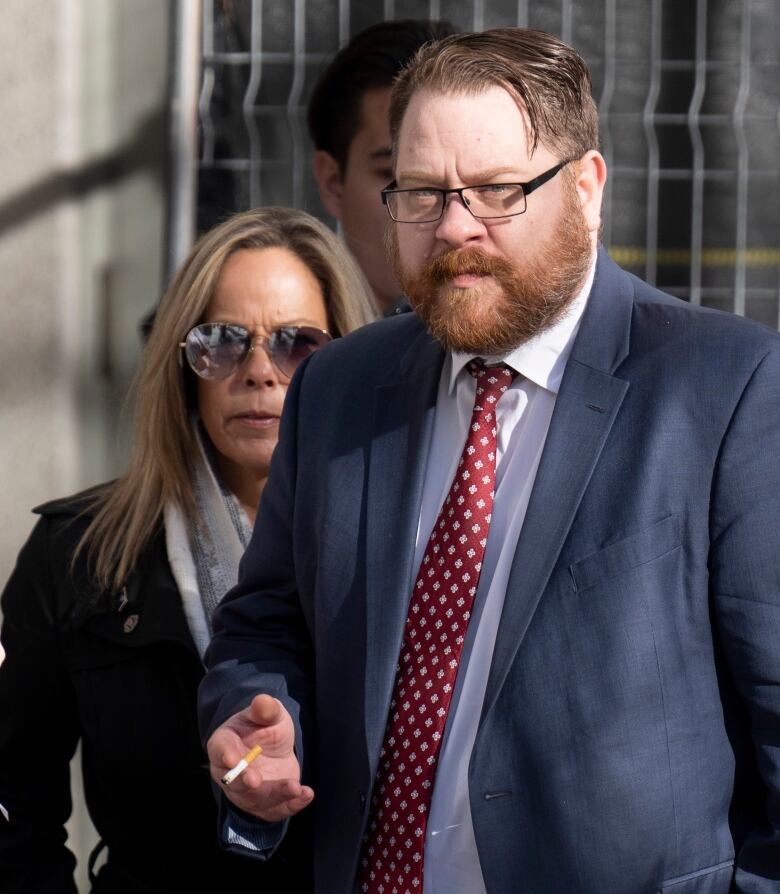 A man wearing a suit and holding a cigarette, with a woman behind him.