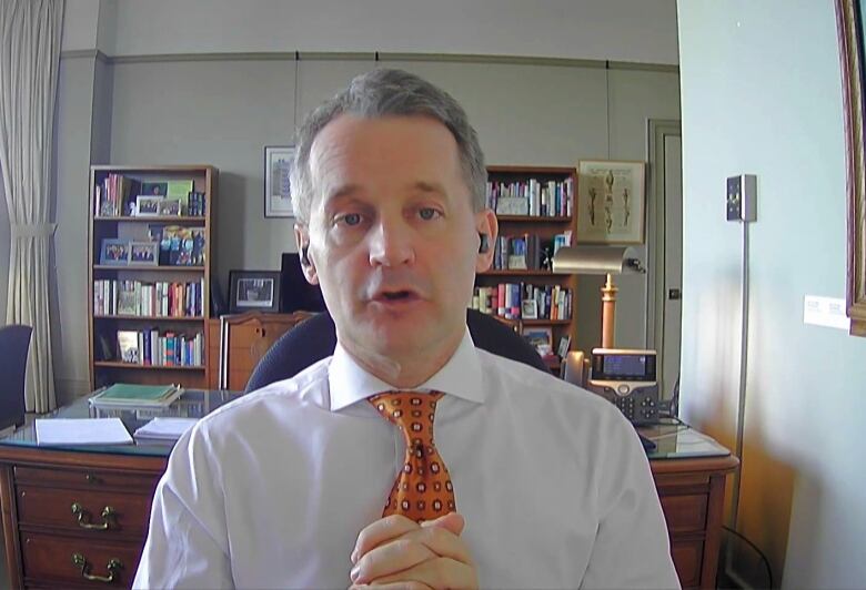 A person wearing a white shirt and orange tie folds his hands while speaking.