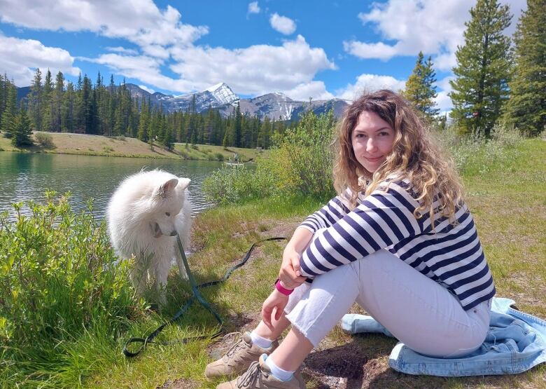 A woman smiles at the camera on a bright, sunny day in the mountains. Her white dog it beside her.