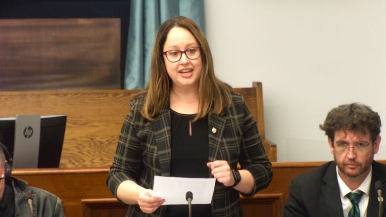 A woman in a blazer with shoulder-length brown hair and holding a paper in front of her speaks into a microphone. 