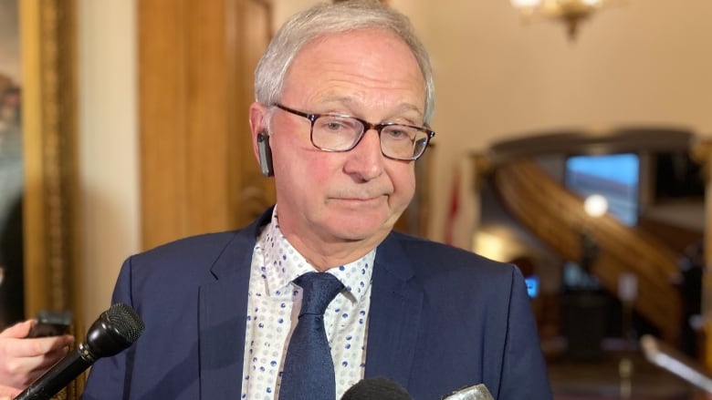 A man in a blue suit and tie shrugs as he answers questions before an array of journalists' microphones. 