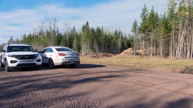 Two RCMP vehicles parked on a dirt road in front of a wooded area.