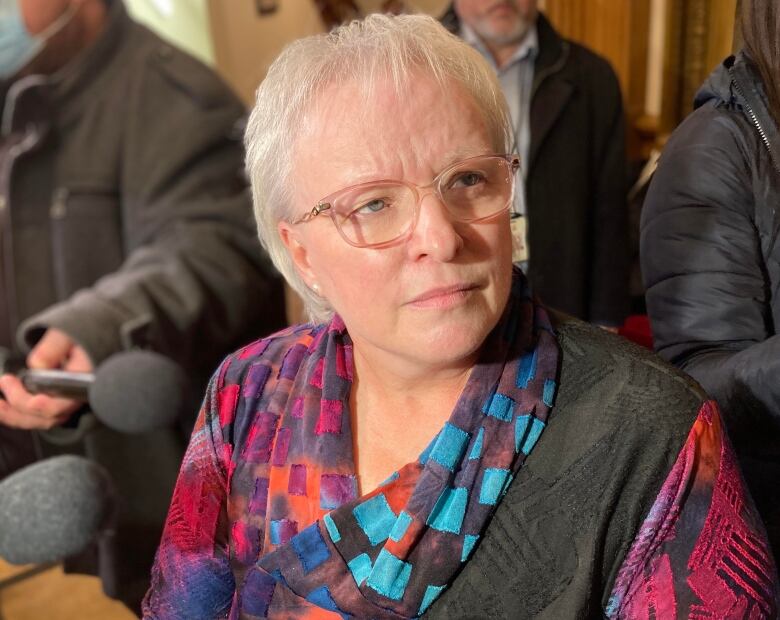 A woman with short white hair and glasses wearing a multi-coloured blouse.