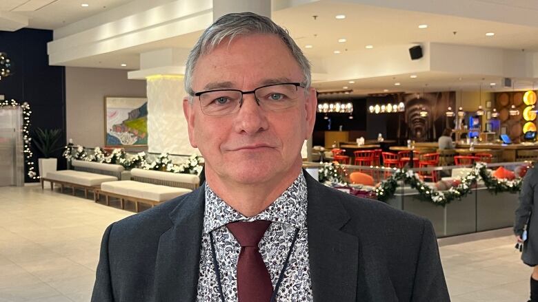 A man with grey hair and glasses stands in a hotel lobby. The lobby is decorated for Christmas.