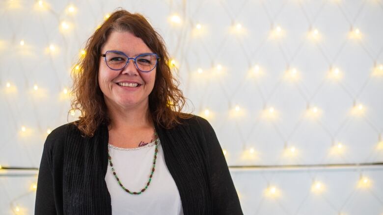 A woman wearing glasses stands before a wall covered in fairies lights.