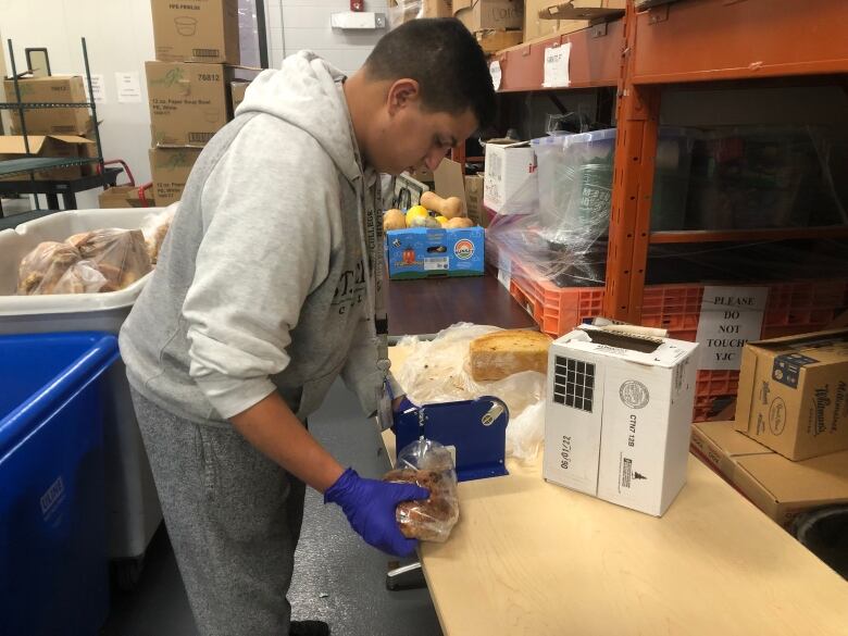 A man wearing a grey hoodie, grey sweatpants and blue latex gloves packages a loaf of bread in a plastic bag