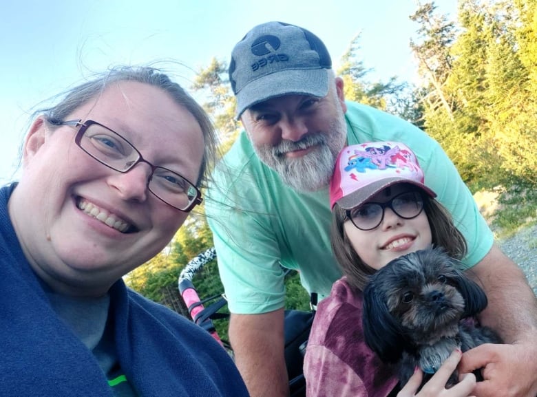 A photo of a woman, left, man, centre, and child, right. They all smile into the camera. On the child's lap sits a small-breed dog.