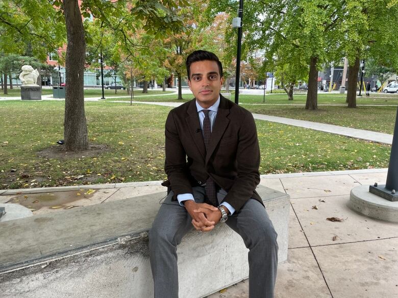 A middle-aged South Asian man wearing a brown blazer sits on a cement block outside. 