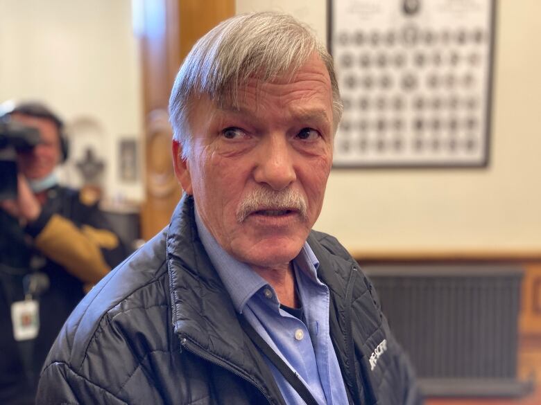 A man wears a dark jacket and light blue shirt. He has a mustache and silver hair.