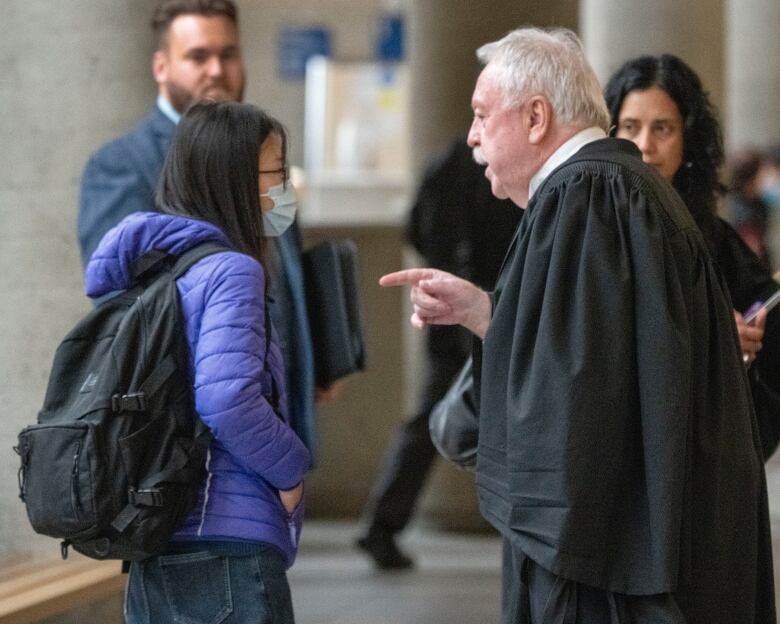 Courthouse hallway with people, a man in black lawyer's robe talks to young woman in blue puffer jacket.