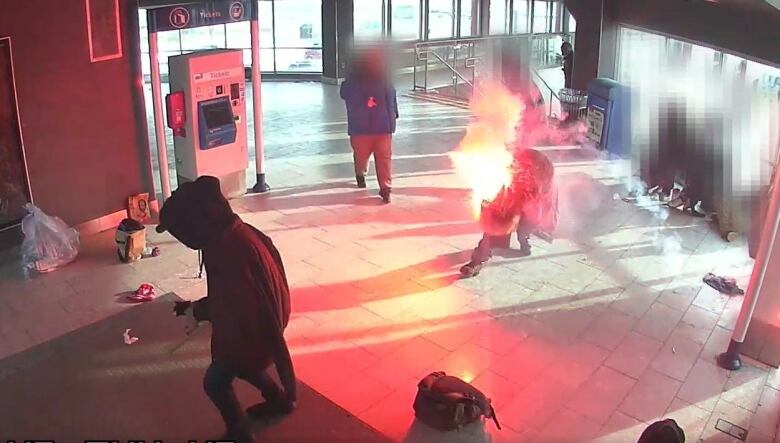 Wide shot inside a Calgary Transit station, with one man in flames. 