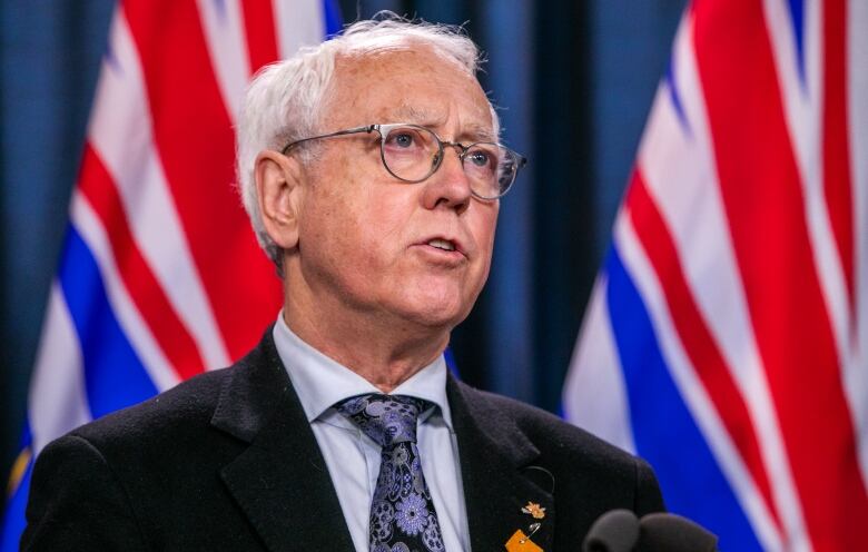 A man with white hair and glasses speaks at a podium in front of B.C. flags.  