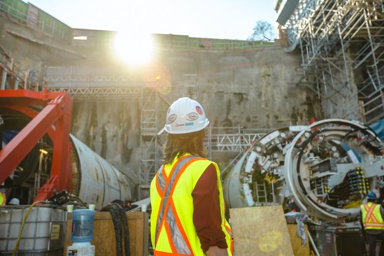 A man in a hard hat and bright construction vest looks on at a tunnel-boring machine in a massive Vancouver construction site.