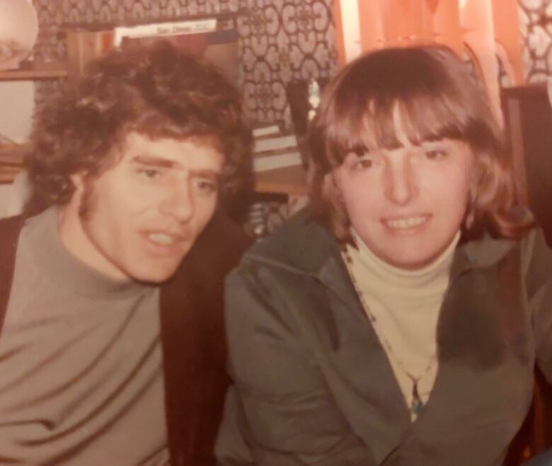 A young man, left, and woman, sit close together and smile. The photo is slightly faded, showing that it was taken decades ago.