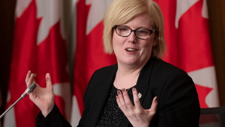 A woman wearing glasses stands in front of a Canadian flag.