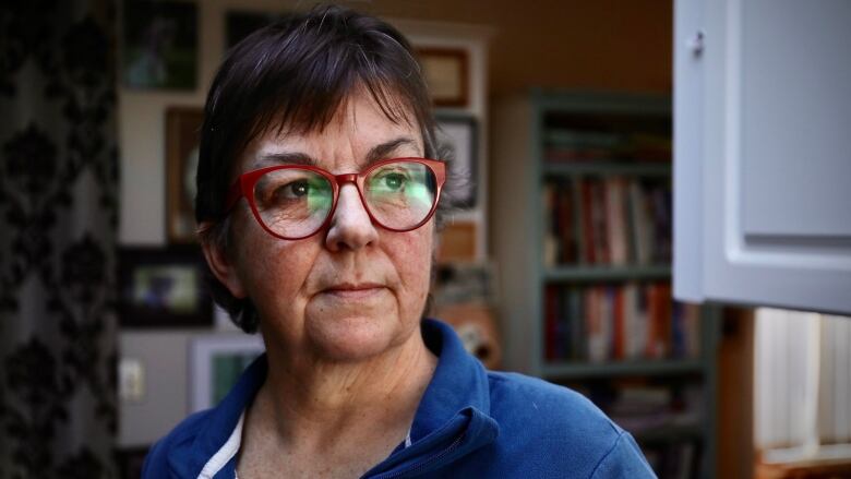 A white woman in her 60s with short brown hair and red-framed glasses stands inside a home. She is looking toward the right of the image.