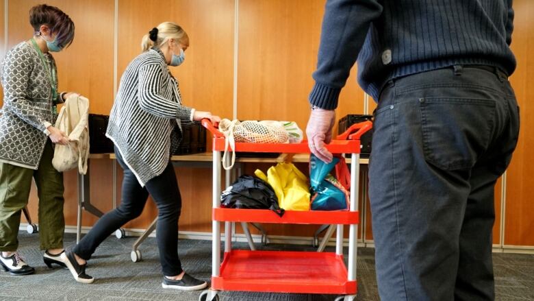 A woman puts food in a cloth bag while a second woman pushes a cart with bags of produce on it.