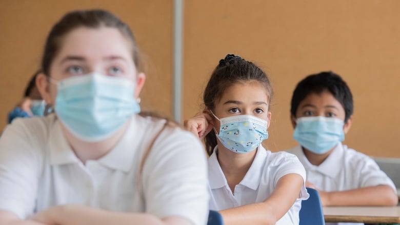 Three students wearing white polo shirts as school uniforms and blue medical face masks sit at desks in a classroom.