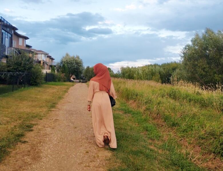 A teen girl wearing a long peach-coloured garment and darker coloured hijab walks along a gravel path in a residential neighbourhood. 