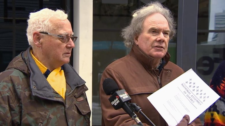 Two men in winter jackets stand outside, speaking to members of the media.