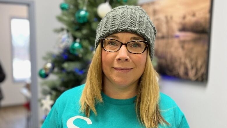 A smiling woman with long blond hair, wearing glasses, a grey knit toque and a turquoise-colored T-shirt, looks into the camera.