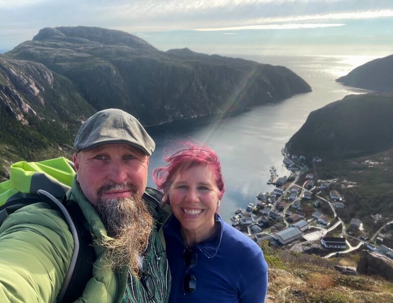 A selfie of a man and woman atop a hill, with a small coastal harbour beneath them. 