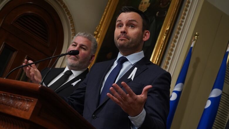 A man at a podium with another man in the background.