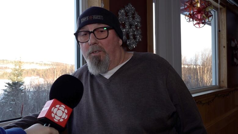 A man wearing a grey sweater and a black hat talks into a microphone.