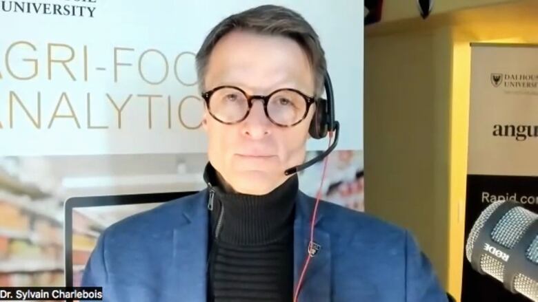 A man wearing a black turtle-neck shirt, a blue suit jacket, glasses and a headset with a microphone