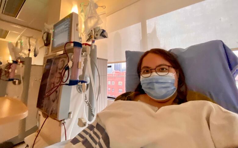 A woman sits up in a hospital bed. To the left there's a machine with tubes. The woman has a neutral expression. She's wearing a mask and is covered by white bedding.