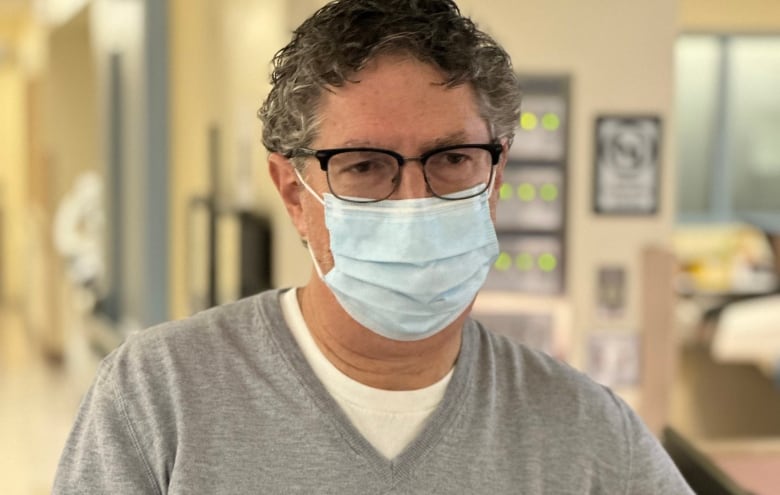 A man in a mask, glasses and  grey sweater stands in a hospital hallway