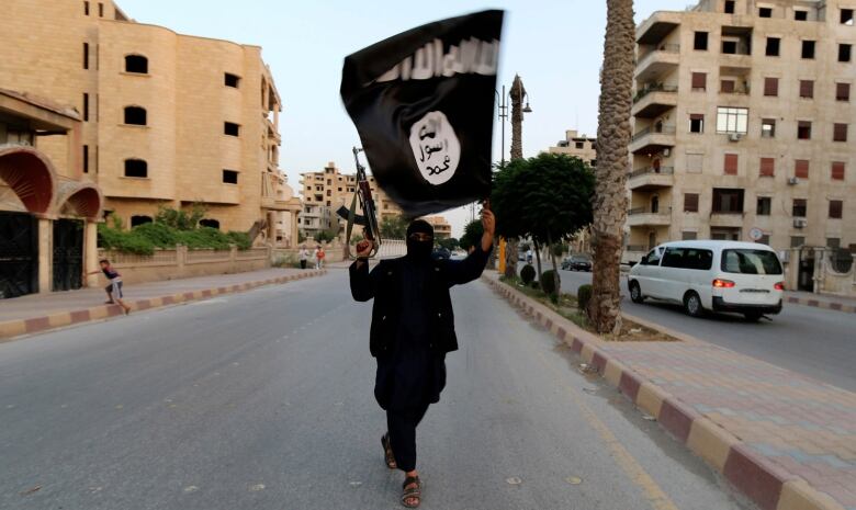 A person dressed all in black with much of their face covered holds a rifle in one hand and a black-and-white Islamic State flag in the other, walking alone in the middle of a street in Raqqa, Syria.