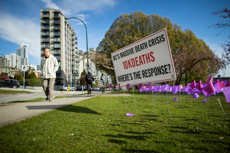 A man walks past a sea of purple flags and a sign that reads 'BC's massive death crisis 10k deaths where's the response?'