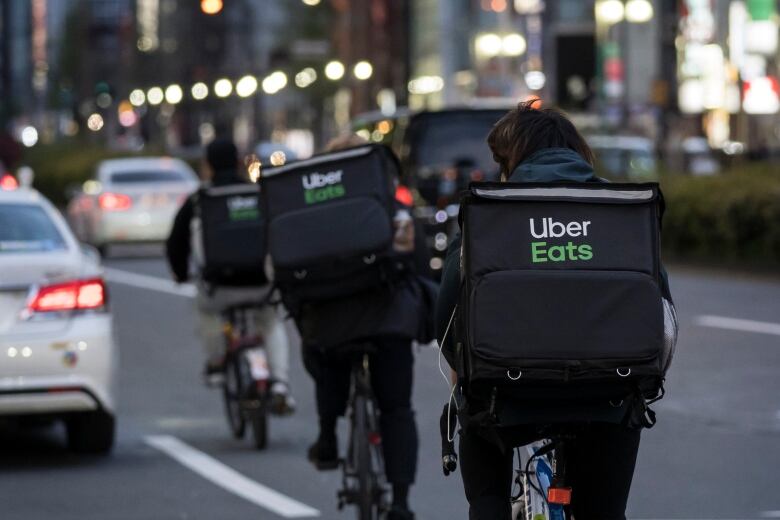 Men ride bikes in the road wearing Uber Eats delivery bags on their backs, 