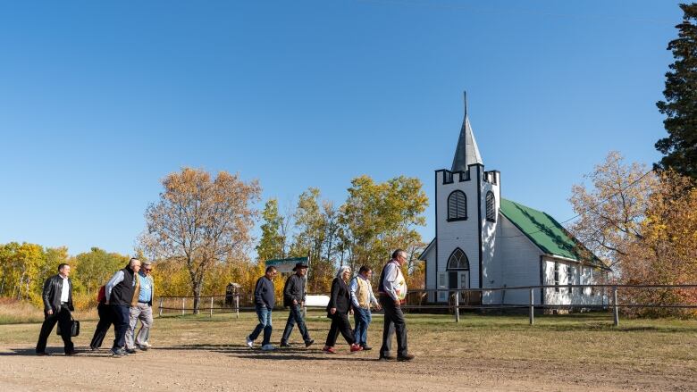 Eight people, including Gov.-Gen. Mary Simon and Indigenous leaders, walk past a church on James Smith Cree Nation.